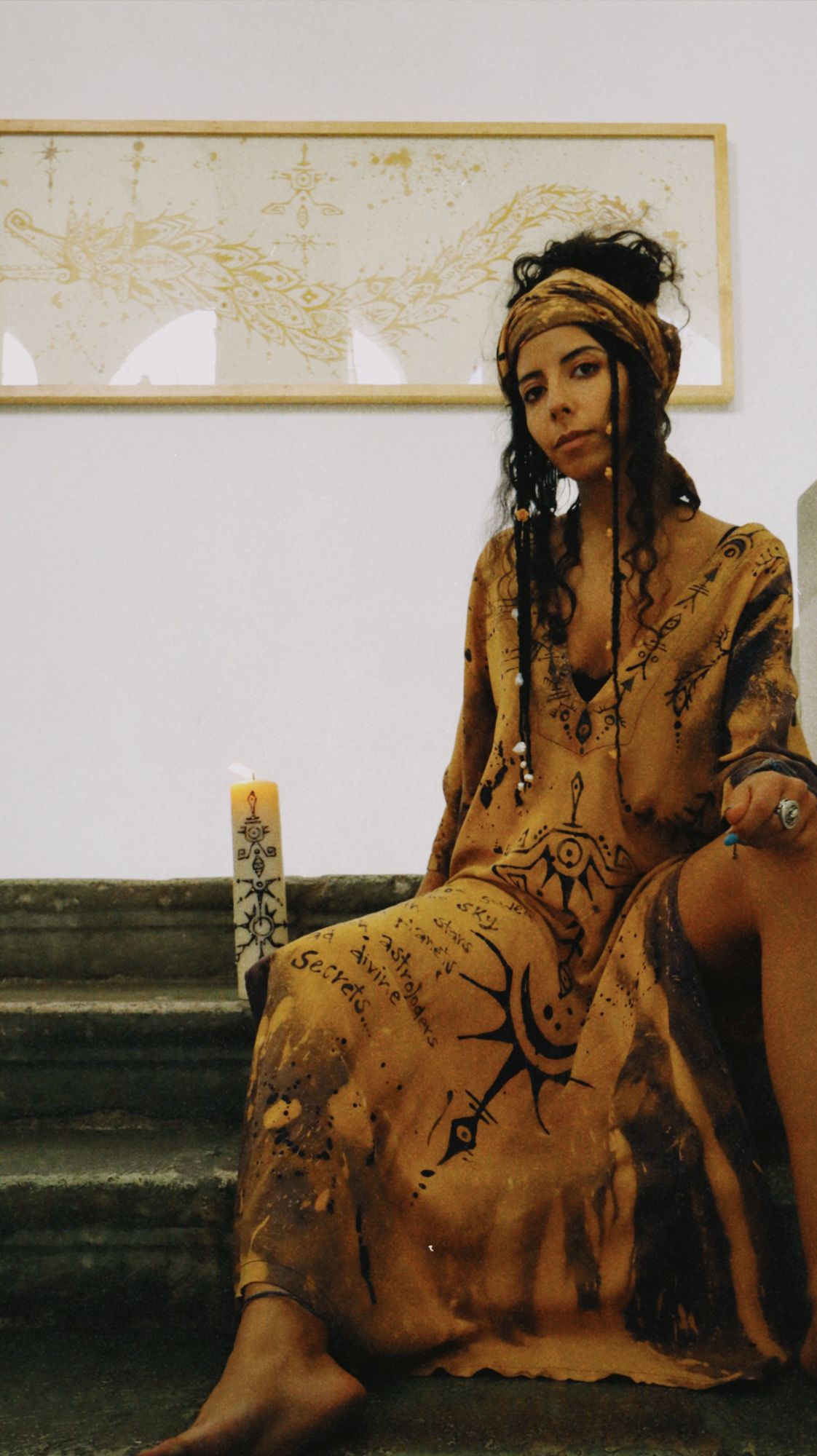Pau seated on stone stairs beside a candle, wearing a ceremonial garment